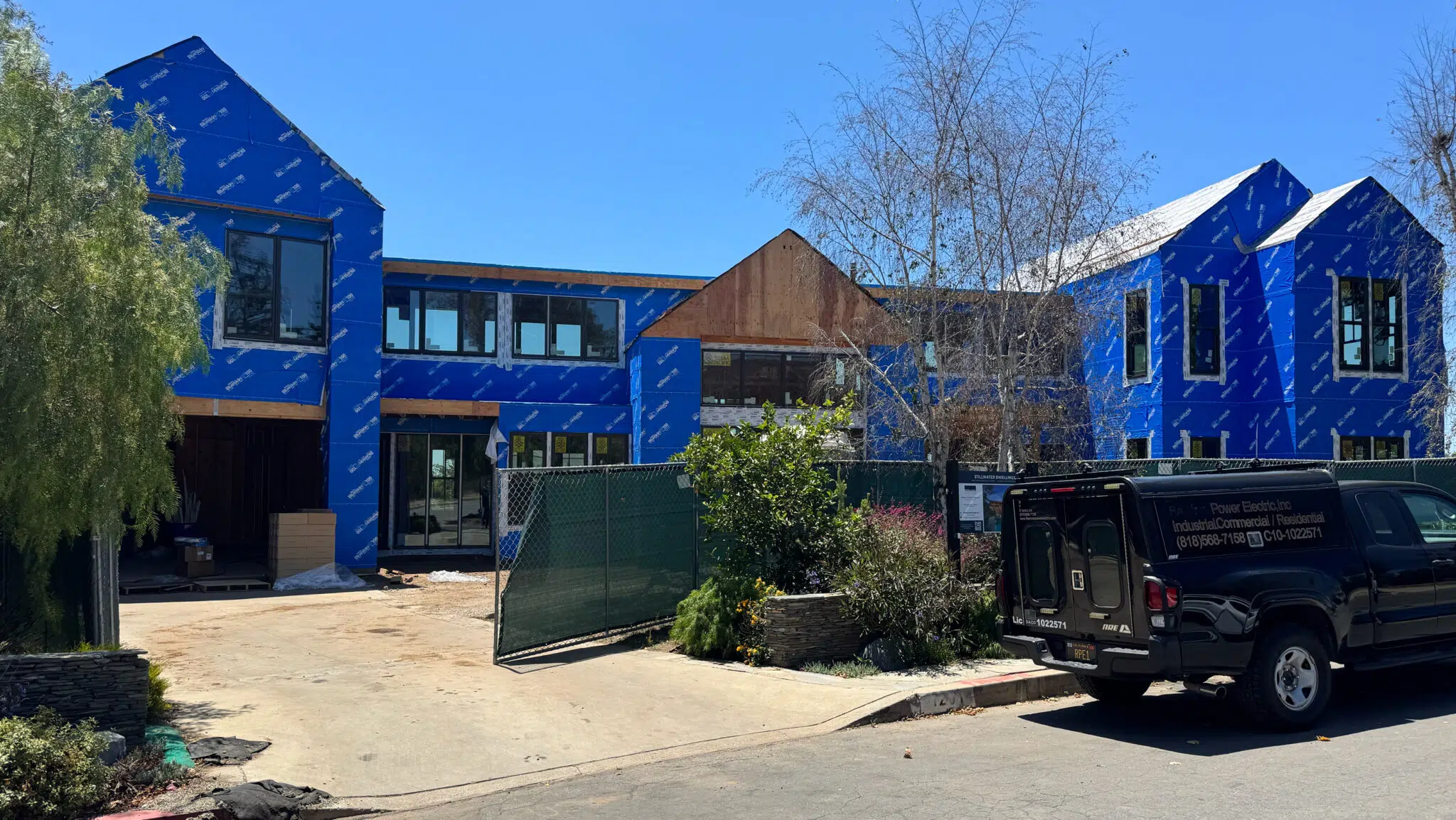 IMG_0230 - Stillwater Dwellings Two-story building under construction wrapped in blue weather barrier, with a black work van in the foreground and a clear blue sky above.