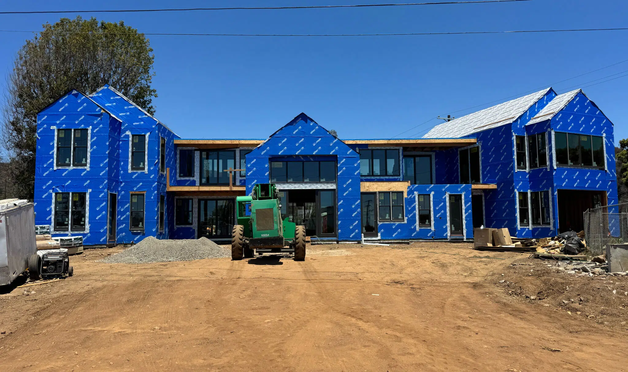 IMG_0235 - Stillwater Dwellings Under-construction row of blue-wrapped houses with large windows on a dirt site, construction equipment in front.
