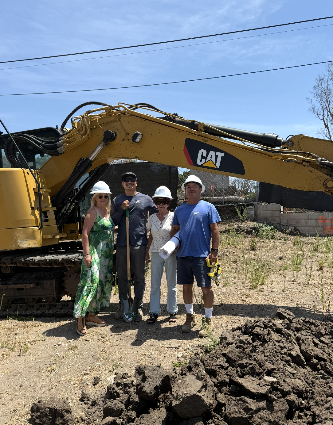 image_0_0 - Stillwater Dwellings Four construction workers wearing hard hats pose beside a large yellow CAT excavator on a dirt site.